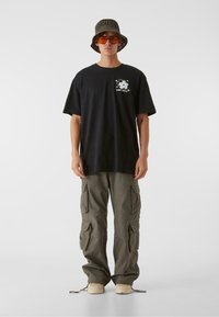 Črna bombažna majica s cvetličnim grafikonom, v kombinaciji z zelenimi cargo hlačami z več žepi in bež športnimi copati. Dodatki vključujejo pleteno klobučko in sončna očala z oranžnim odtenkom.