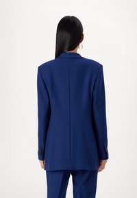 Marinblå skräddarsydd kavaj i ett slätt tyg, med notchad krage, tvåknappsmanschetter och en rak skärning med en enkel bakre slits.