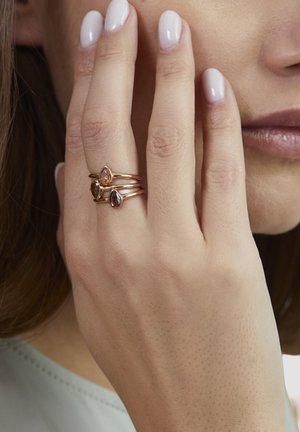 Three stacked rings in rose gold feature pear-shaped, colorful gemstones. The hand displays smooth skin and neatly manicured nails.