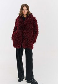 Žena v tmavě červeném huňatém kožichu, černých kalhotách s širokými nohavicemi a černých mokasínách stojí na světlém jednobarevném pozadí.