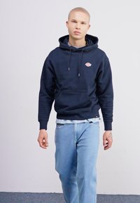 Felpa blu navy con tasca frontale e logo, abbinata a jeans azzurri. Il tessuto sembra morbido; la vestibilità è casual e rilassata.