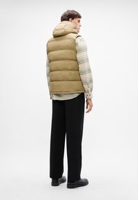 Gilet in piuma khaki con cappuccio, caratterizzato da cuciture orizzontali, indossato sopra una maglietta a righe. Abbinato a pantaloni neri e stivali.