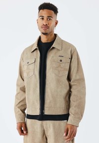 Chaqueta con cremallera de color beige con dos bolsillos en el pecho. Presenta una tela texturizada y costuras en forma de estrella en los bolsillos. Se lleva sobre una camiseta negra.