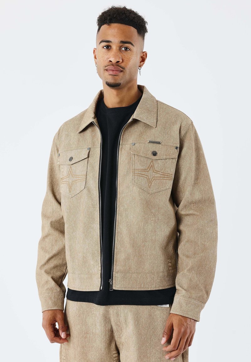 Chaqueta con cremallera de color beige con dos bolsillos en el pecho. Presenta una tela texturizada y costuras en forma de estrella en los bolsillos. Se lleva sobre una camiseta negra.