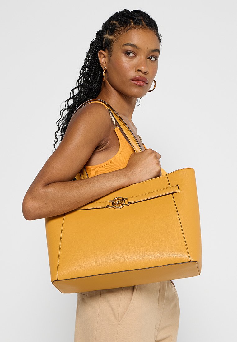 Femme avec des cheveux tressés portant un débardeur orange et un pantalon beige tient un grand sac à main jaune moutarde avec un logo circulaire doré.