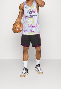 Camiseta de baloncesto con un patrón de tie-dye en tonos pasteles, logotipo negro en el frente, combinada con pantalones cortos oscuros con acentos púrpuras contrastantes y zapatillas Nike.