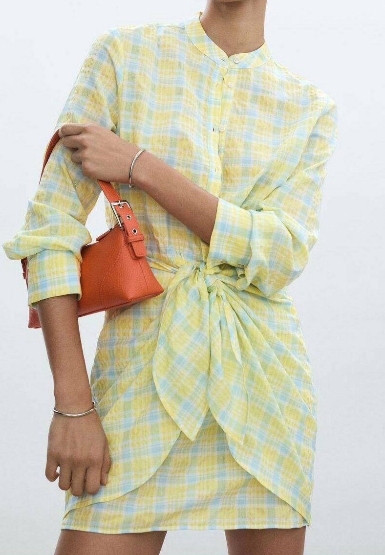 Personne portant une robe-chemise à carreaux jaune et bleue avec un nœud à l'avant, tenant un petit sac à main orange sur un bras, avec des bracelets argentés aux deux poignets.