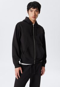 Joven vestido con una sudadera negra con cremallera y pantalones negros a juego, de pie contra un fondo claro y liso.