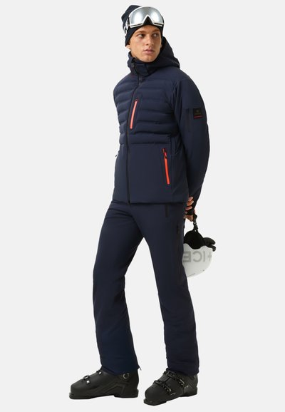 Doudoune marine avec des accents orange vif, poches zippées et capuche, associée à un pantalon de ski assorti et des bottes de ski noires.