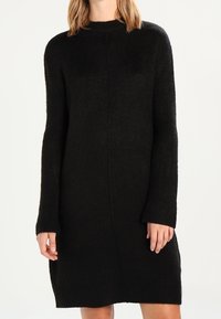 Donna che indossa un vestito maglione nero a maniche lunghe, lunghezza ginocchio, su uno sfondo bianco semplice.