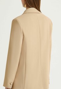 Blazer beige con texture liscia, spalle strutturate e colletto a incavo. Presenta un dettaglio con polsini a due bottoni e una vestibilità sartoriale.