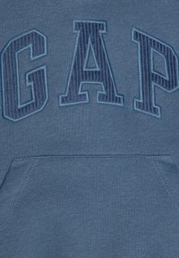 Hoodie en polaire bleue avec un grand logo "GAP" brodé en bleu plus foncé. Comprend une poche kangourou et un tissu doux et texturé.
