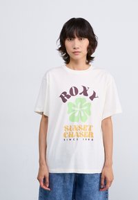 Hvit bomull T-skjorte med korte ermer, med et grønt blomsterdesign og fet tekst i brunt og oransje, kombinert med blå denimbukser.