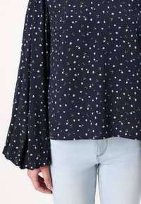 Blusa navy a fiori con maniche lunghe a campana. Presenta piccoli fiori rosa e bianchi, foglie verdi e un tessuto leggero. Abbinata a jeans chiari.