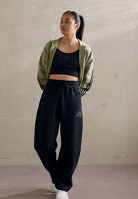 Sportinė apranga, susidedanti iš šviesiai žalios zip-up džemperio, juodos maikutės, ir laisvų juodų sportinių kelnių su pilkų logotipų akcentais. Baltos sportinės avalynės.