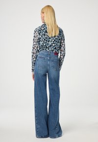 Bloemenblouse met lange mouwen van doorzichtig materiaal, gecombineerd met hoge taille, wijduitlopende blauwe jeans met achterzakken en een rood labeldetail.