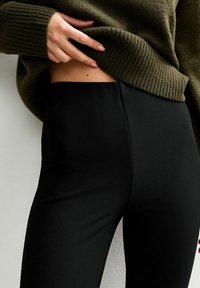 Persoon in zwarte aansluitende broek en een groene geribbelde trui, die de zoom optilt om een deel van hun blote taille te onthullen.