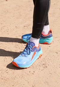 Scarpe da trail running blu e arancioni con una parte superiore in mesh traspirante, suola testurizzata e accenti riflettenti, posizionate su una superficie sabbiosa.