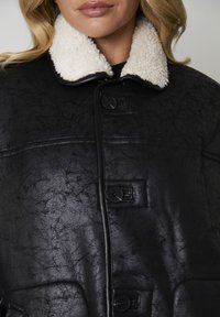 Sort imiteret læderjakke med en krémfarvet shearlingkrave, der har knaplukninger og en tekstureret overflade. Detaljeret syning og synlige lommer.