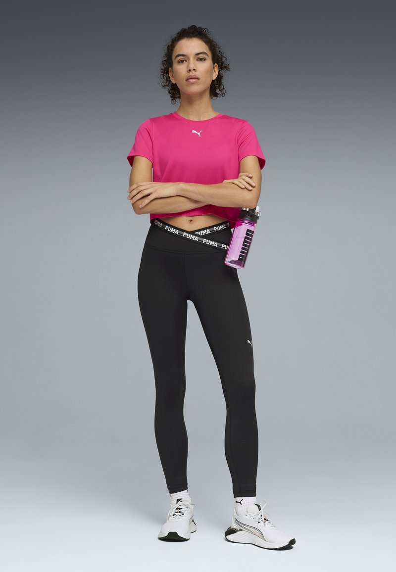 Pink athletic t-shirt, cropped design, PUMA logo. Black leggings with branded waistband. White athletic shoes. Water bottle in hand.