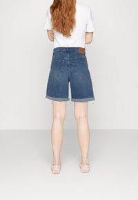 Personne aux longs cheveux roux portant un short en jean retroussé, un t-shirt blanc et des chaussures rose clair, debout les bras croisés, dos à la caméra.