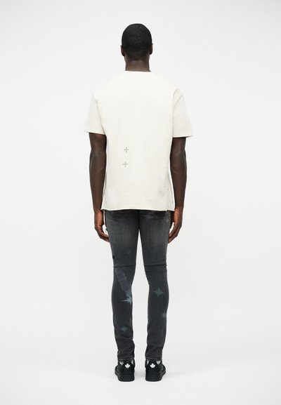 Άνδρας που στέκεται πλάτη, φορώντας ένα απλό λευκό κοντομάνικο πουκάμισο, ξεβαμμένο στενό μαύρο τζιν με σχέδια αστεριών και μαύρα παπούτσια.