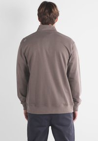 Veste pull marron avec un col montant, des manches longues et un ourlet côtelé. Elle présente un petit logo sur l'épaule gauche. Tissu doux et texturé.