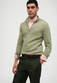 Grön zip-up tröja i mjukt material, tillsammans med en ljusrosa skjorta med knappar. Mörkgröna byxor och flätat brunt bälte.