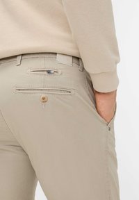 Beige Baumwollhosen mit glatter Textur, ausgestattet mit zwei Gesäßtaschen mit horizontalen Öffnungen und einem Knopfdetail.