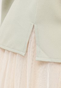 Detailní záběr na světle zelenou texturovanou látku s bočním rozparkem, který odhaluje světle růžovou průhlednou síťovinu pod ní.