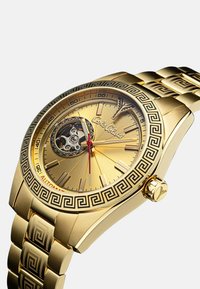 Goldene Uhr mit einem runden Zifferblatt, kunstvollen Mustern auf der Lünette und dem Armband, einer sichtbaren mechanischen Bewegung und roten Akzenten für Stunde- und Minutenzeiger.