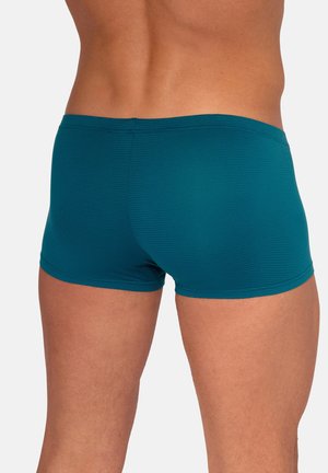 Mann trägt enge türkisblaue Badehosen, gezeigt von hinten, die die strukturierte Stoffoberfläche und die Passform an den Oberschenkeln und der Taille hervorheben.