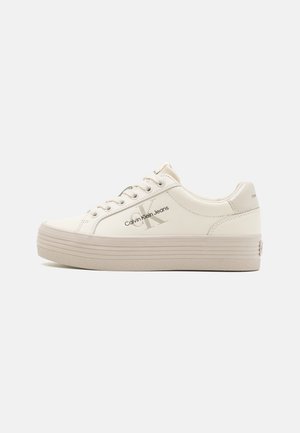 Sneaker i lav platform cremefarvet med hvide snørebånd og Calvin Klein Jeans-logo på siden mod en ensfarvet baggrund.