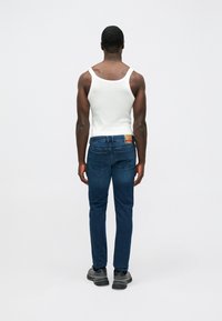 Μπλε τζιν παντελόνι με �ίσια γραμμή, συνδυασμένο με λευκό αμάνικο μπλουζάκι. Σημαντικές λεπτομέρειες περιλαμβάνουν τη ραφή των τσεπών και μια επώνυμη δερμάτινη ετικέτα.