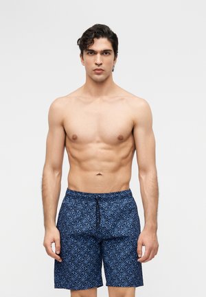 Homme torse nu aux cheveux bouclés foncés portant un short de bain bleu avec un motif géométrique bleu clair couvrant l'ensemble, debout devant un fond uni.