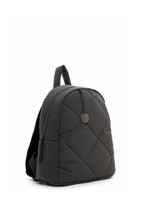 Schwarzer gesteppter Rucksack mit Frontreißverschluss und kleinem rundem Metallschild, das an einer Kette hängt, aufrecht stehend vor weißem Hintergrund.