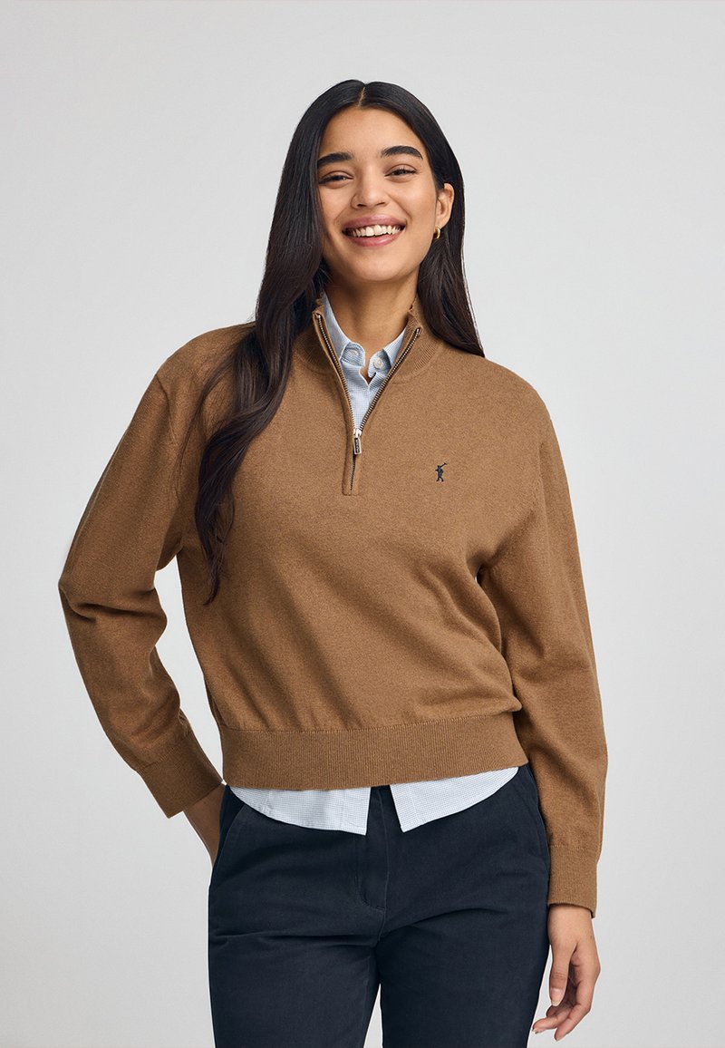 Maglione marrone con zip realizzato in un tessuto morbido; presenta un orlo e polsini a coste, abbinato a una camicia colletto azzurro sotto.