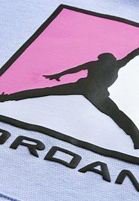 Silhouette di un giocatore di basket che salta in nero su uno sfondo triangolare rosa e bianco con parte del testo "JORDAN" sottostante su tessuto.