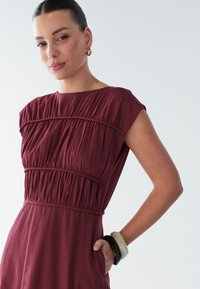 Fialovoružové šaty s nariasenými detailmi okolo korzetu, krátkymi rukávmi, okrúhlym výstrihom a pohodlným strihom, doplnené o náramkové náramky.