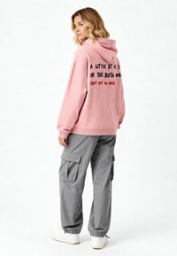 Hoodie oversize rosa con cappuccio, caratterizzato da testo nero e rosso sul retro. Abbinato a pantaloni cargo grigi e sneaker bianche.