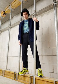 Jeune homme portant un bob, une veste bleu marine, un t-shirt tie-dye, un pantalon à fines rayures et des baskets jaune néon, debout sur une plateforme en bois tenant des poteaux en métal.