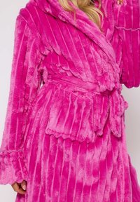 Fuchsia pluche badjas met verticale strepen, grote kap, uitlopende mouwen en een strikceintuur. Bevat een opgestikte zak aan de voorkant. Zachte textuur.