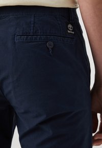 Navy blue trousers with a buttoned back pocket, featuring a stitched logo patch. Smooth fabric with a casual, relaxed fit.