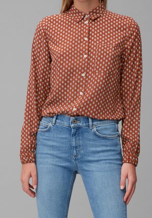 Kobieta w rdzawej, zapinanej na guziki koszuli z długim rękawem i białym geometrycznym wzorem oraz jasnobłękitnych, wysokich dżinsach na tle szarego tła.