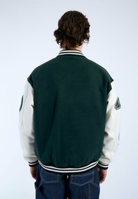 Giacca varsity verde e crema con colletto a maglia, maniche bicolore e dettagli ricamati. Presenta una superficie morbida e orlo a coste.