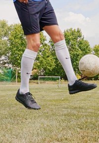 Schwarze Fußballschuhe, weiße knielange Socken und schwarze Shorts, getragen von einer Person auf Gras, die einen Fußball unter einem klaren Himmel tritt.