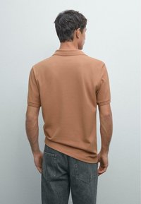 Man facing backward wearing a short-sleeve camel polo shirt and dark gray jeans against a plain light gray background.