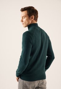 Dunkelgrüner Pullover mit hohem Kragen, ausgestattet mit strukturierten Strickmustern auf den Schultern und einem glatten Körper, kombiniert mit hellgrauen Hosen.