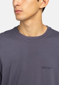 Graues Rundhalsausschnitt-T-Shirt aus Baumwolle. Besitzt ein kleines schwarzes Logo auf der Brust. Glatte Textur mit kurzen Ärmeln. Unisex-Passform.