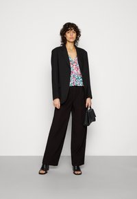 Blazer oversized preto sobre uma blusa de mangas largas vibrante com padrão floral, combinado com calças largas pretas e sandálias de salto bloco. Pequena bolsa preta.